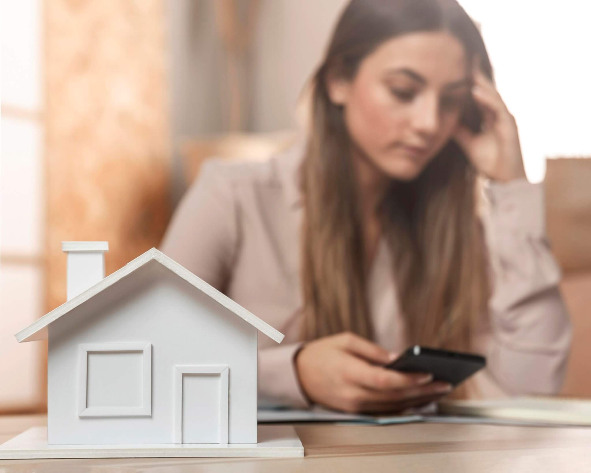 Mujer preocupada revisando su teléfono mientras sostiene documentos, con una maqueta de casa en primer plano